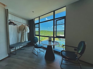 A room with a large window, a round table, two chairs, a bench, and a clothing rack with garments.