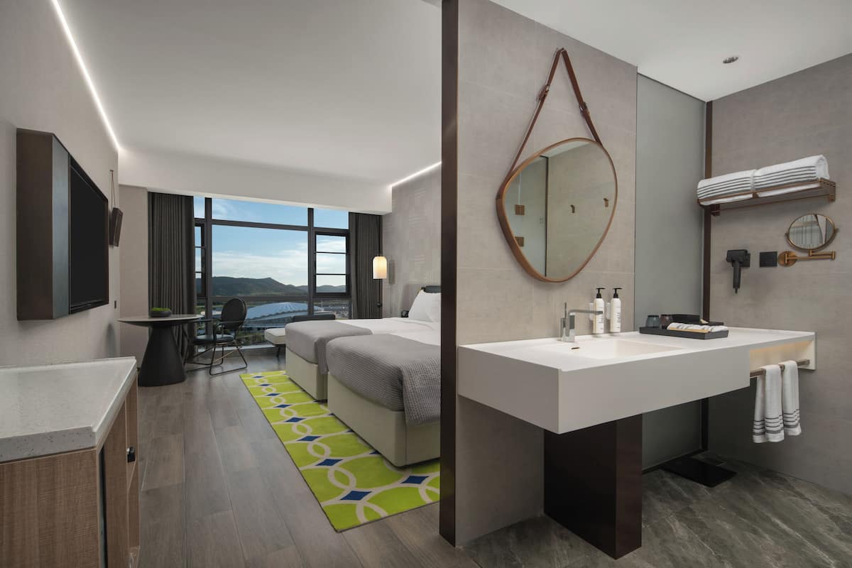 Une chambre d’hôtel moderne avec un grand lit, une salle de bain dotée d’un grand miroir et d’un lavabo, et une vue sur les montagnes.