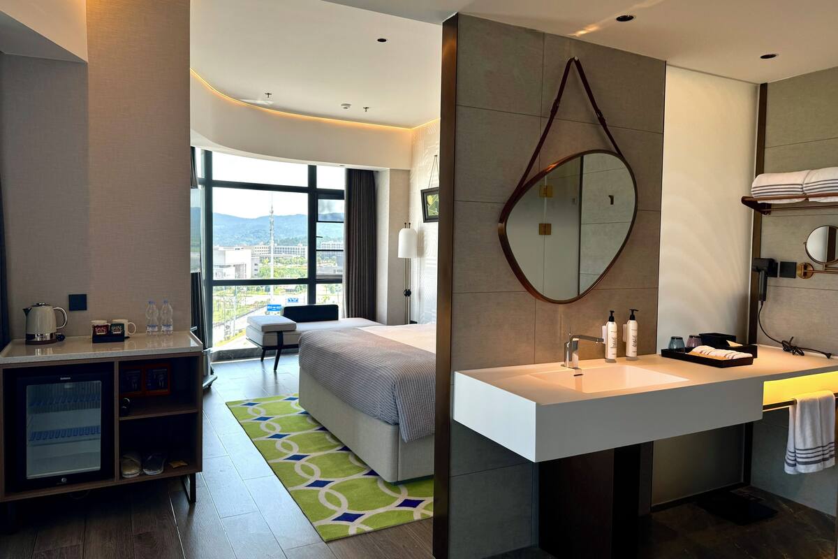 Une chambre d’hôtel moderne équipée d’un lit, d’un bureau avec une bouilloire, d’une grande fenêtre offrant une vue, d’une salle de bain avec un miroir et un lavabo.