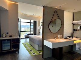 Una habitación de hotel moderna con cama, escritorio con hervidor, ventana grande con vistas, baño con espejo y lavamanos.