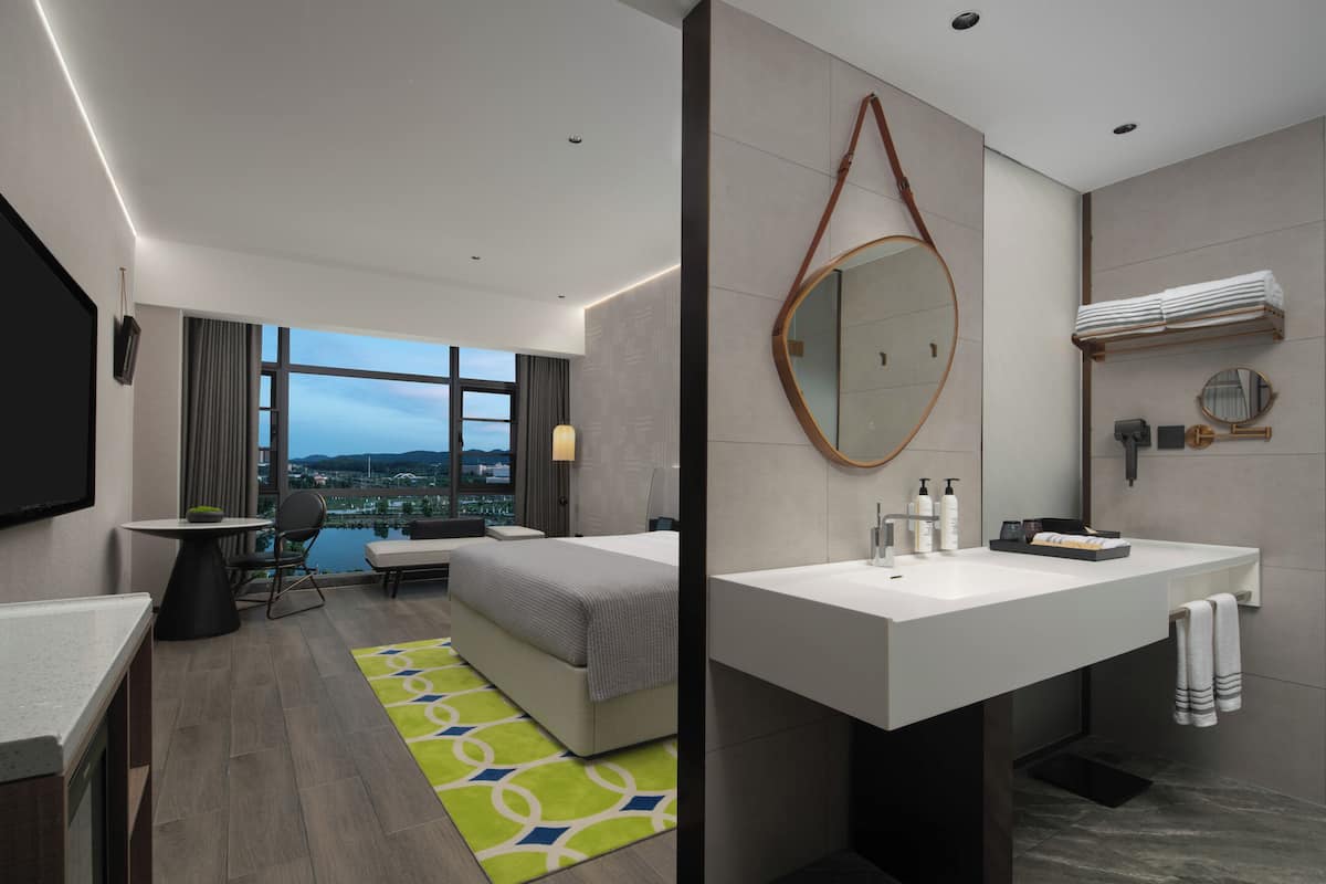 Une chambre d’hôtel moderne comprenant un lit, un bureau, une grande fenêtre avec vue, une salle de bain avec un lavabo et un miroir, ainsi qu’une petite table avec une chaise.