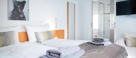 A hotel room with two beds, a large mirror, a television mounted on the wall, and a glass shower enclosure.