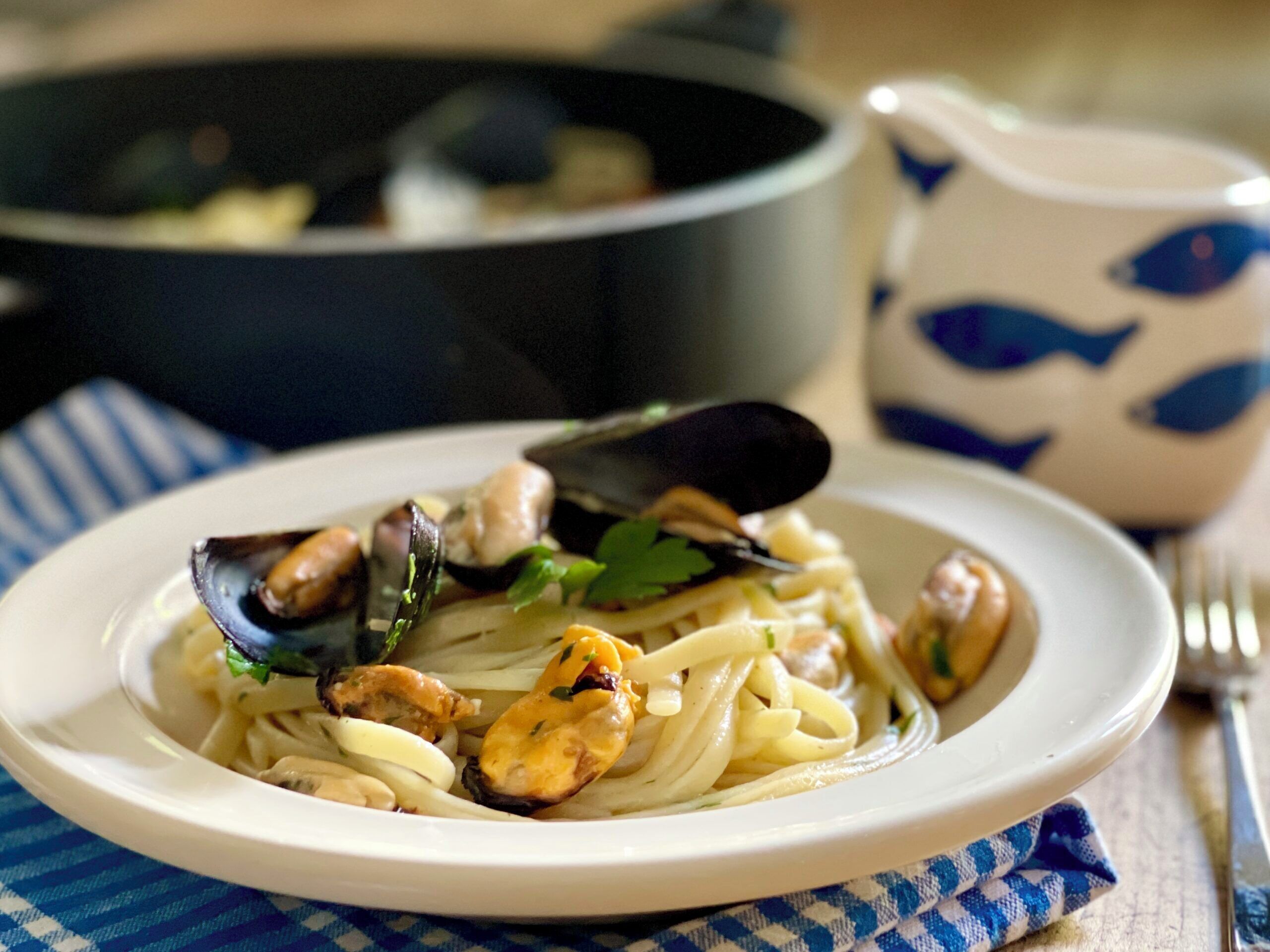 A plate of pasta with mussels and chicken, a pot in the background, and a blue and white patterned napkin.