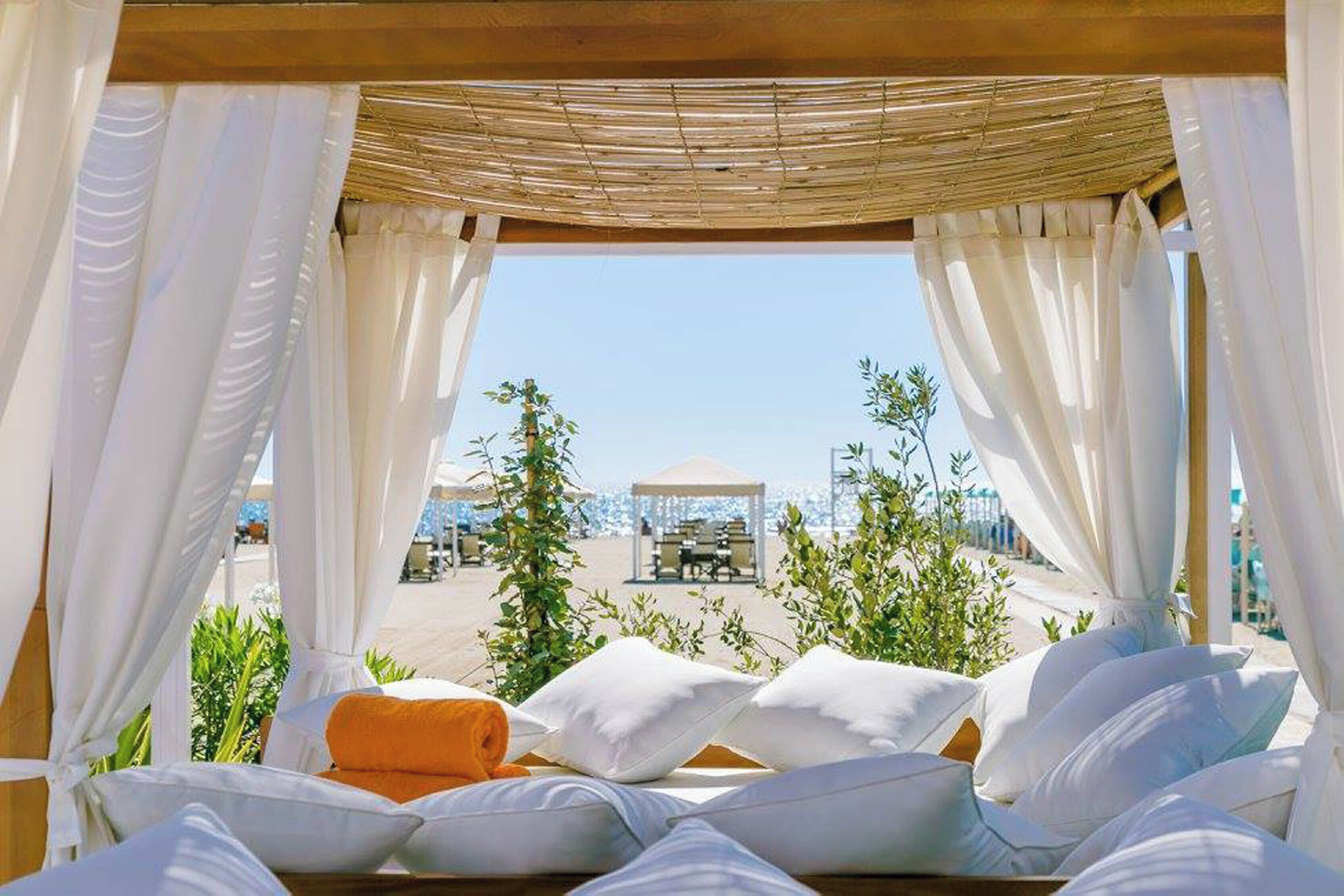 Ein sonnendurchflutetes Zimmer mit einem Bett, weißen Vorhängen und Blick auf Strand und Sonnenschirme.