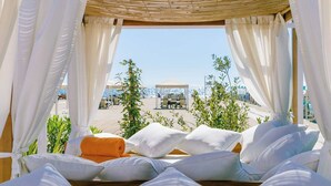 Een zonnige kamer met een bed, witte gordijnen en uitzicht op een strand en parasols.