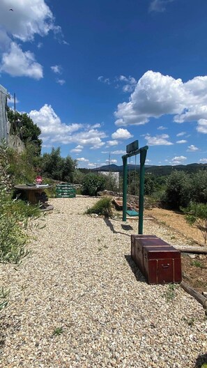 A gravel path leading to a green swing set and a red suitcase in a rural setting with a clear sky and scattered clouds.