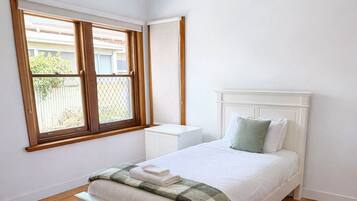 A bedroom with a wooden floor, a white bed with a green and white blanket, a white nightstand, and a window with wooden frames.