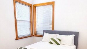 A neatly made bed with a grey headboard, a striped blanket, and a pillow. There are two windows with wooden frames and blinds. - 2BR Seaside Retreat in Largs North (Largs North)