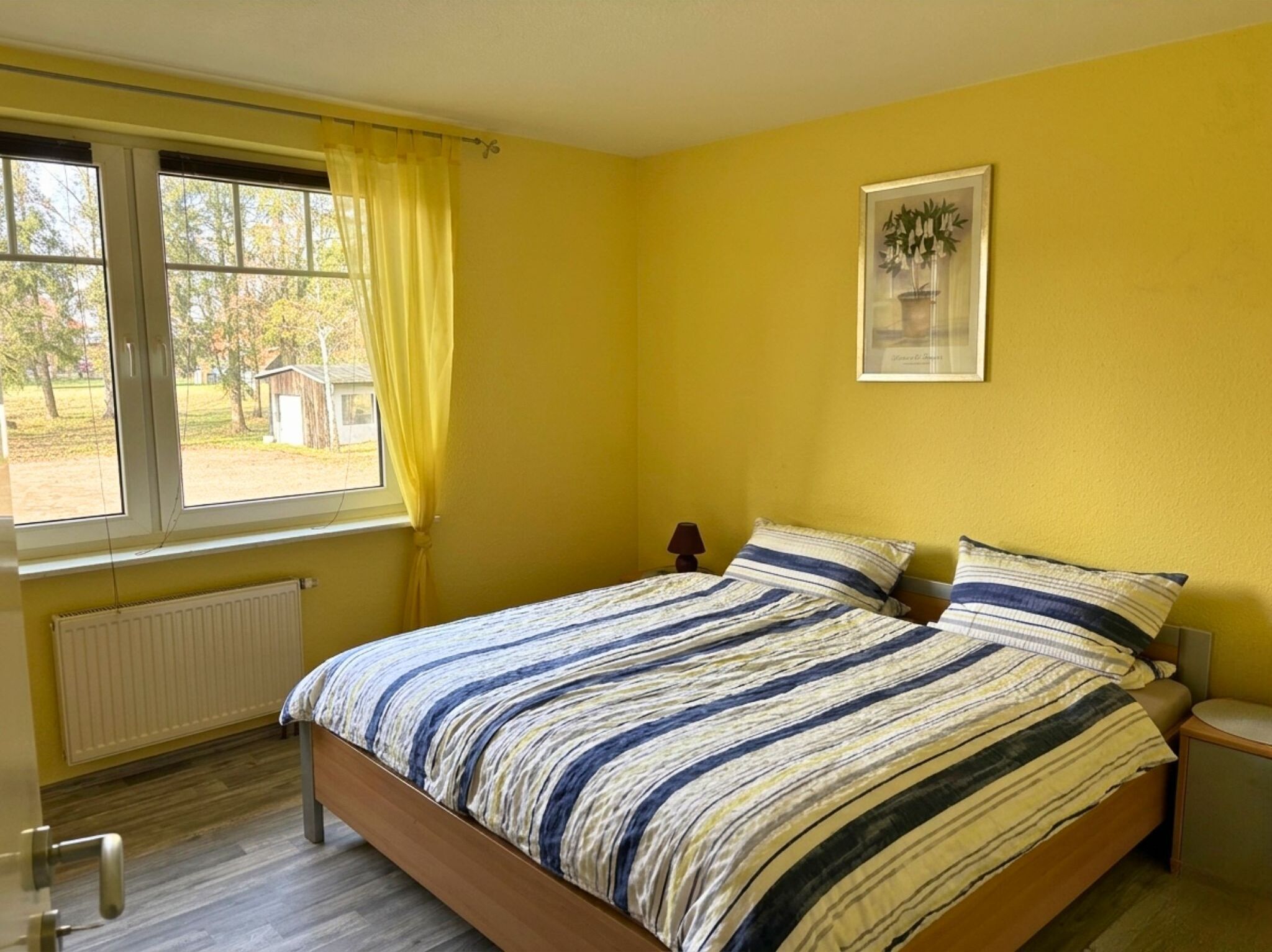 Helles und freundliches Schlafzimmer in einem Apartment auf Möllers Morgen. Die Wände sind in einem warmen Gelbton gehalten, was dem Raum eine einladende und gemütliche Atmosphäre verleiht. Das Zimmer ist ausgestattet mit einem Doppelbett, das mit blaugrauen, gestreiften Bettdecken bezogen ist.