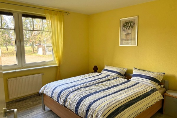 Helles und freundliches Schlafzimmer in einem Apartment auf Möllers Morgen. Die Wände sind in einem warmen Gelbton gehalten, was dem Raum eine einladende und gemütliche Atmosphäre verleiht. Das Zimmer ist ausgestattet mit einem Doppelbett, das mit blaugrauen, gestreiften Bettdecken bezogen ist.