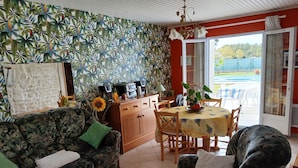 A living room with a patterned sofa, a coffee table with remote controls, a dining area with a table and chairs, and a view through a glass door. - Au Calme 4 star house 2 bedrooms large swimming pool 6x12 m sea and town 1,5 km (Saint-Pierre-d'Oléron)