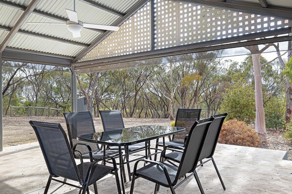 Un patio exterior con una mesa de vidrio y sillas de metal, rodeado por una pérgola de metal y árboles.