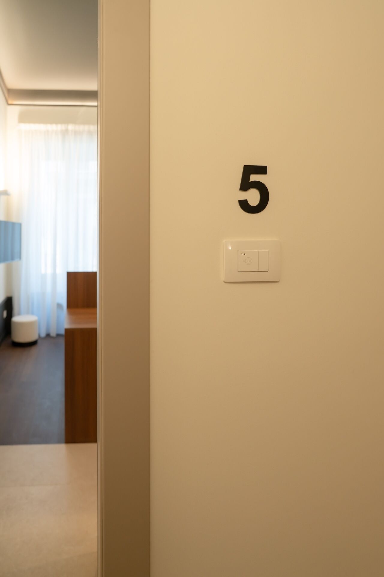 A hotel room with a door labeled '5', a light switch, and a glimpse of a bathroom with a toilet and a sink.