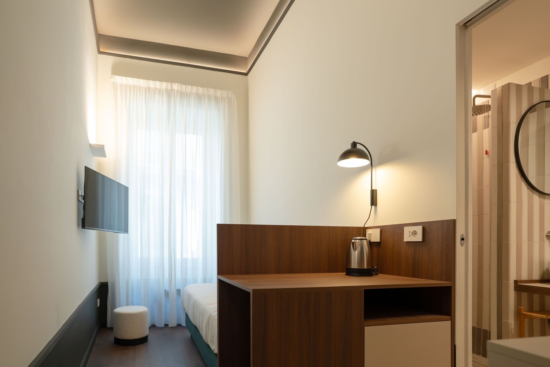 A hotel room with a bed, a desk, a chair, a TV, a lamp, and a shower area.