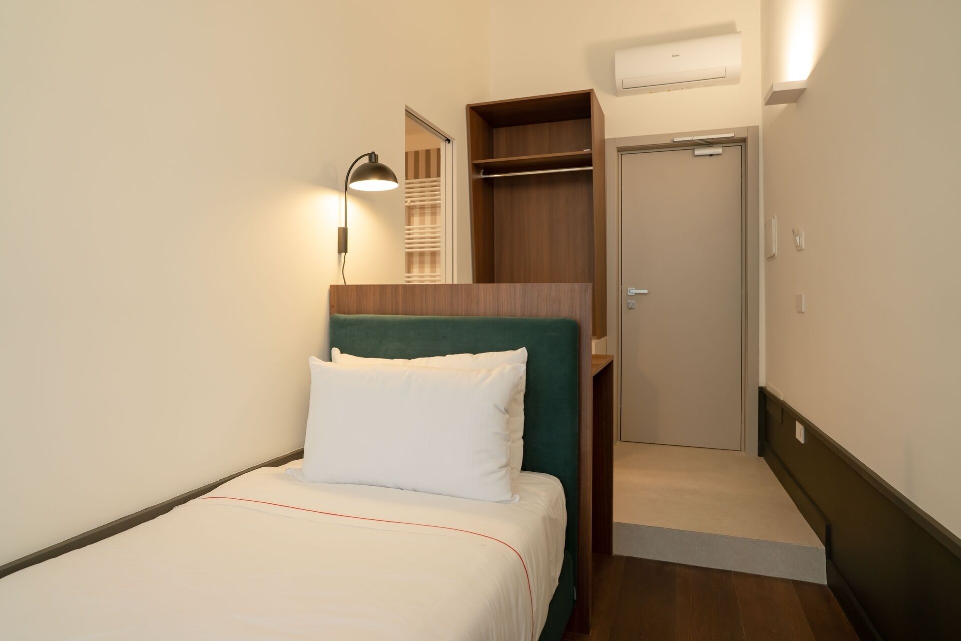 A hotel room with a bed, a bedside lamp, a wardrobe, and an air conditioning unit.
