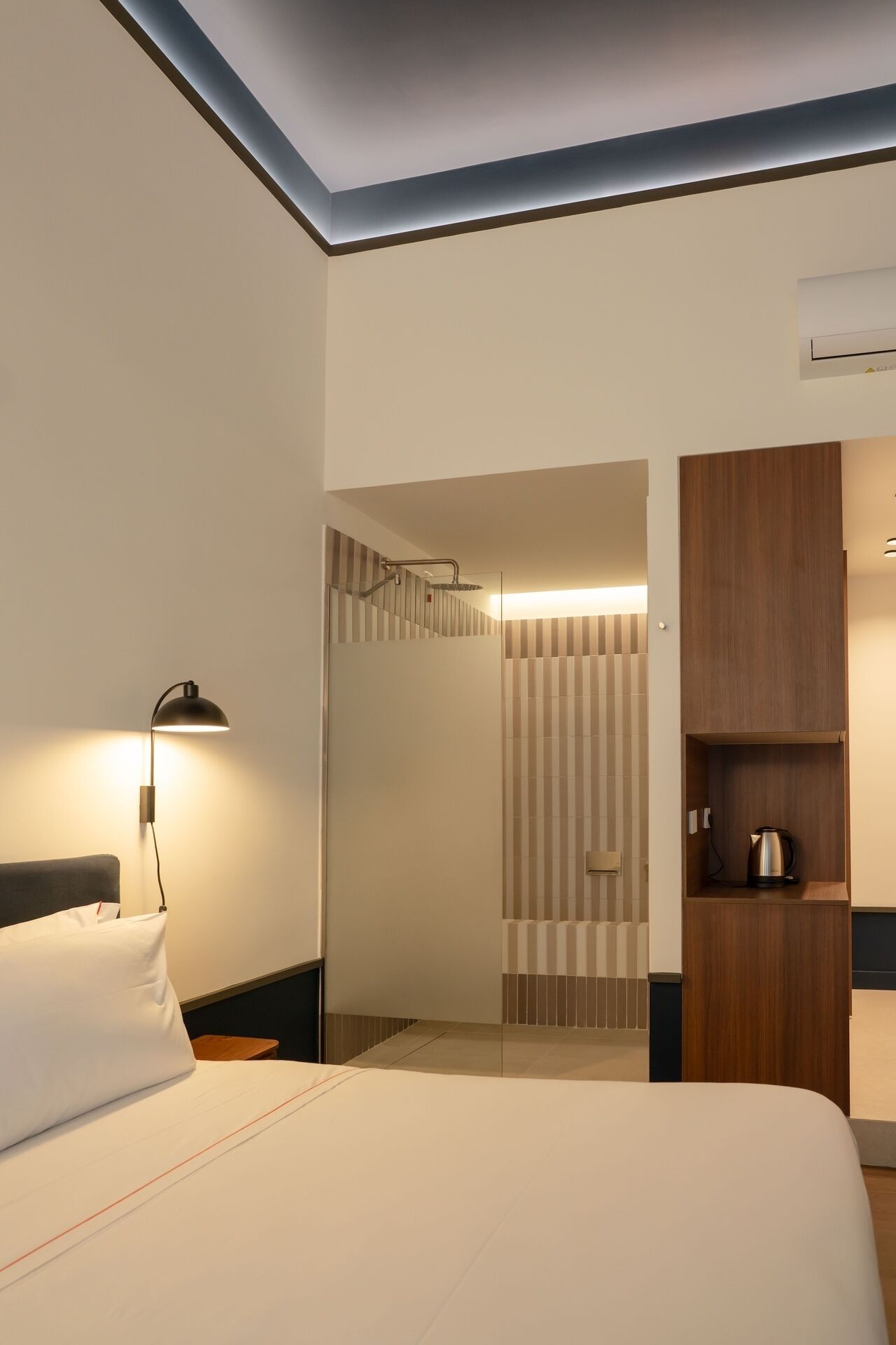 Una camera d'albergo con un letto, un comodino, una lampada, un armadietto in legno e un angolo doccia.