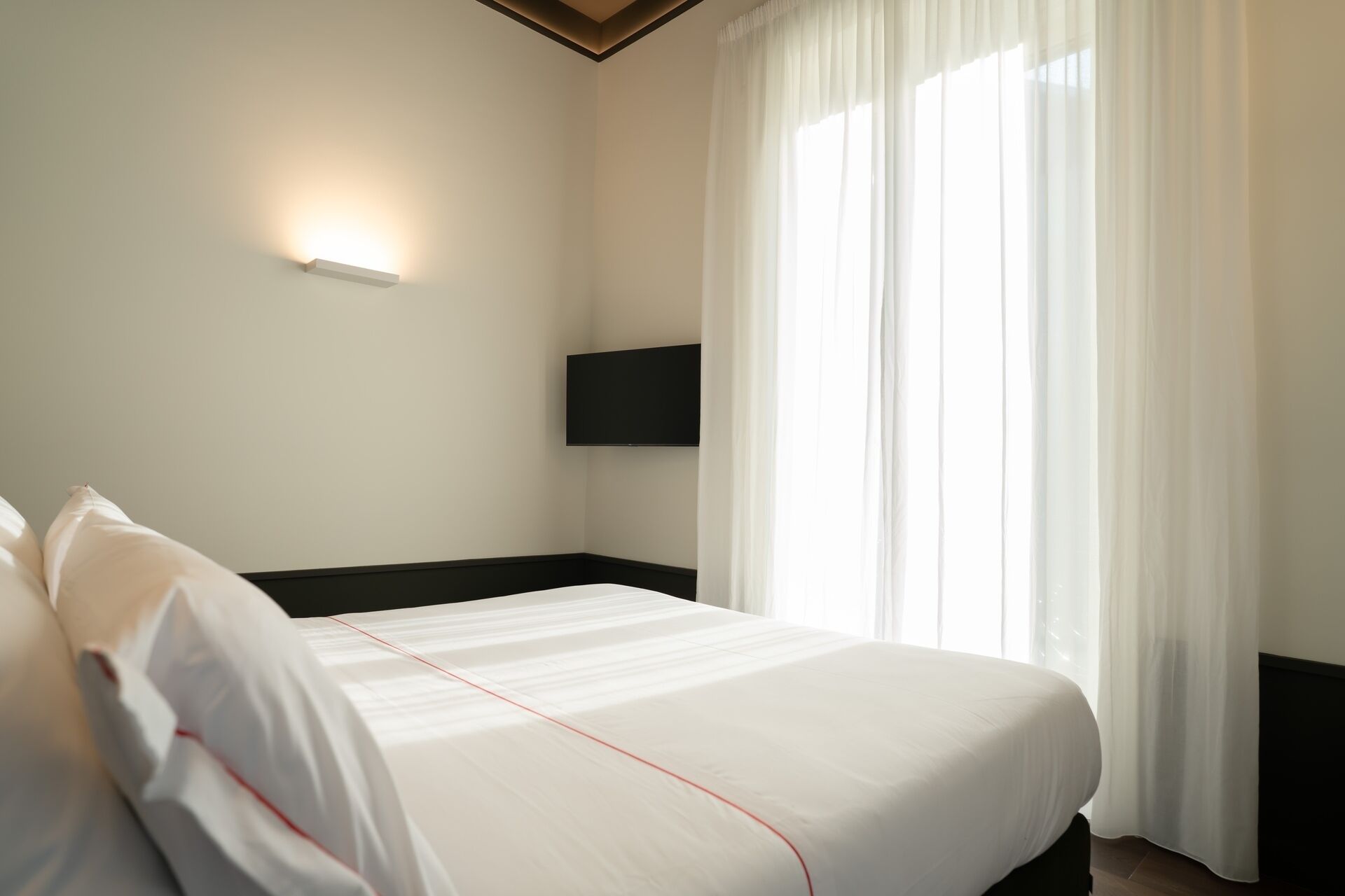 A hotel room with a bed, a flat-screen TV mounted on the wall, and a large window with sheer curtains.