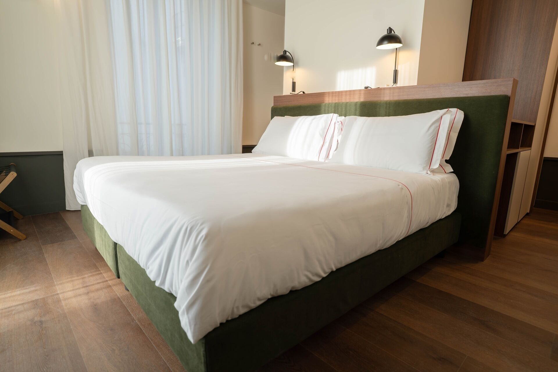 Un letto rifatto con biancheria bianca e un poggiatesta verde, due abat-jour e un pavimento in legno.