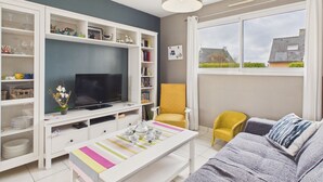A living room with a TV, white shelves, a yellow chair, and a window with a view of houses. - Beach 200m away, house for 6 with garden (Guissény)