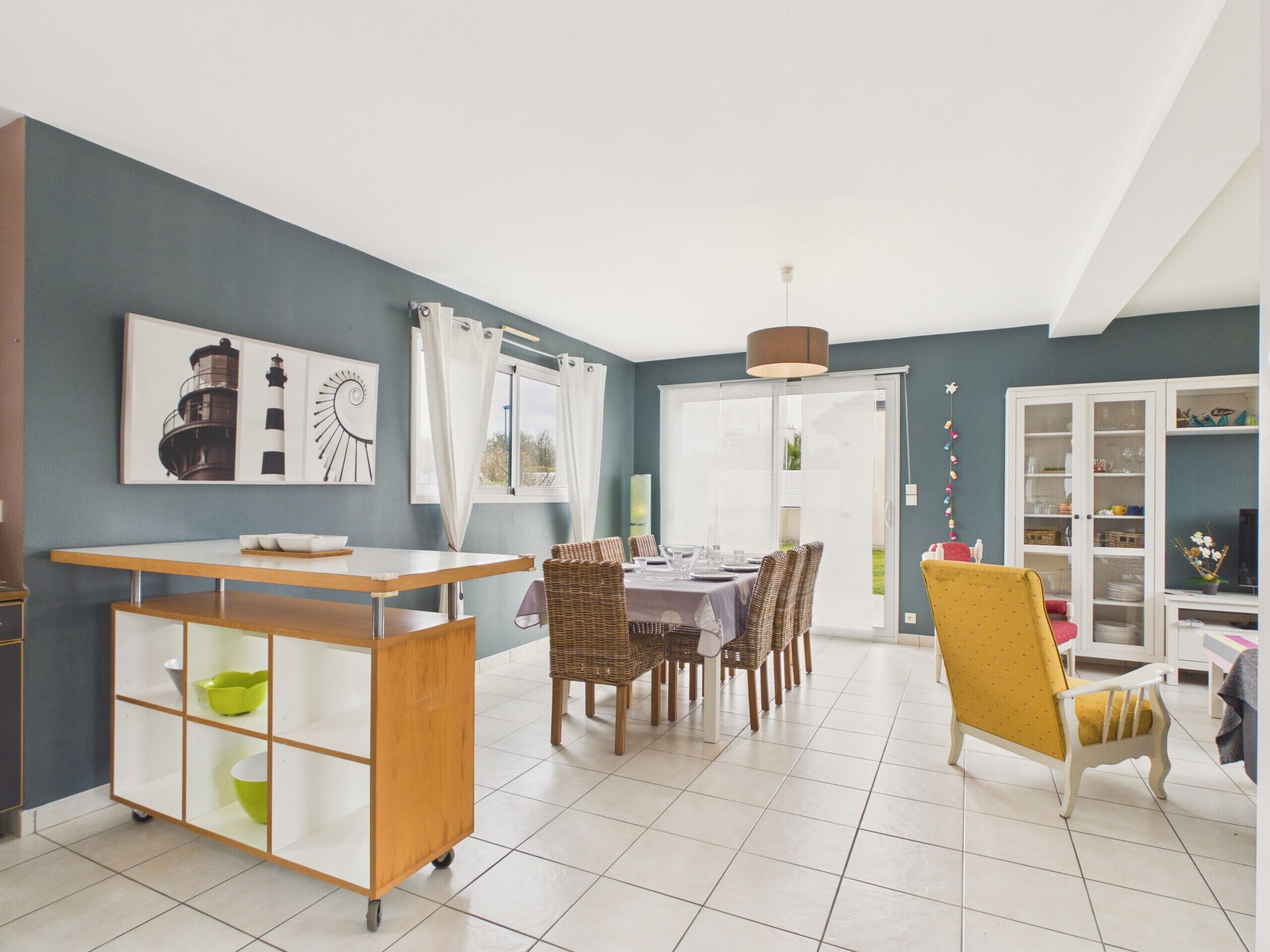 Un coin repas avec une table dressée pour quatre personnes, entourée de chaises en osier. Un petit chariot surmonté d’une vitre est placé devant la table.