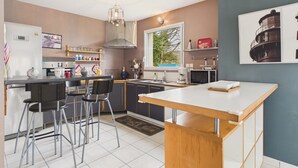 A modern kitchen with a central island, bar stools, a built-in oven, and a window with a view of greenery. - Beach 200m away, house for 6 with garden (Guissény)