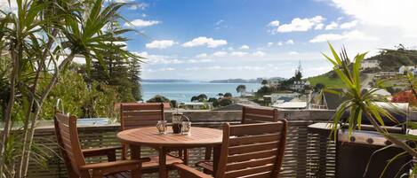 Ein Holztisch mit Stühlen auf einer Terrasse, mit Blick auf das Meer und eine ferne Stadtlandschaft.