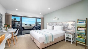 A modern hotel room with a large bed, a desk with chairs, a small bedside table, and a magazine rack.