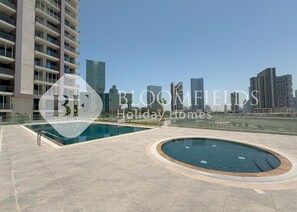 Ein modernes Wohngebäude mit Poolbereich auf dem Dach, das einen großen rechteckigen Pool und einen kleineren runden Pool vor einer Skyline der Stadt zeigt.