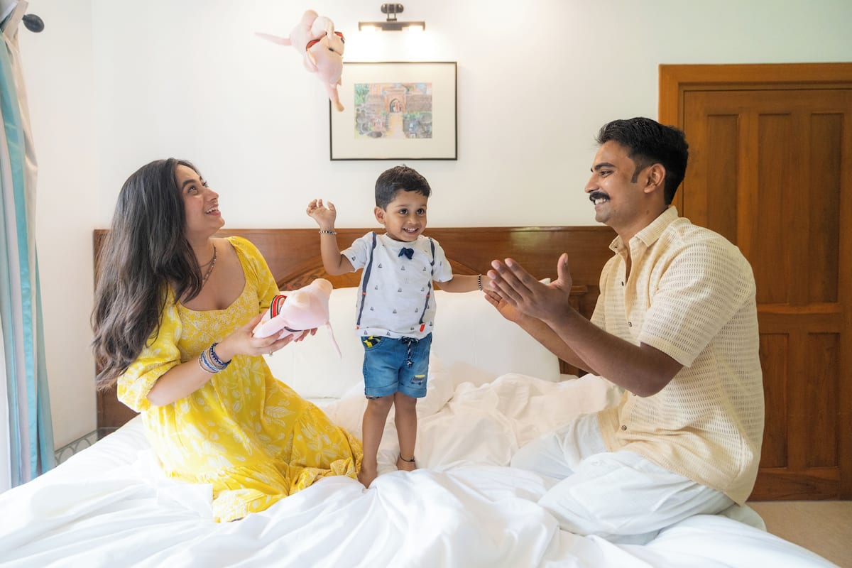 A family on a bed with a wooden headboard, a framed picture on the wall, and a door in the background.