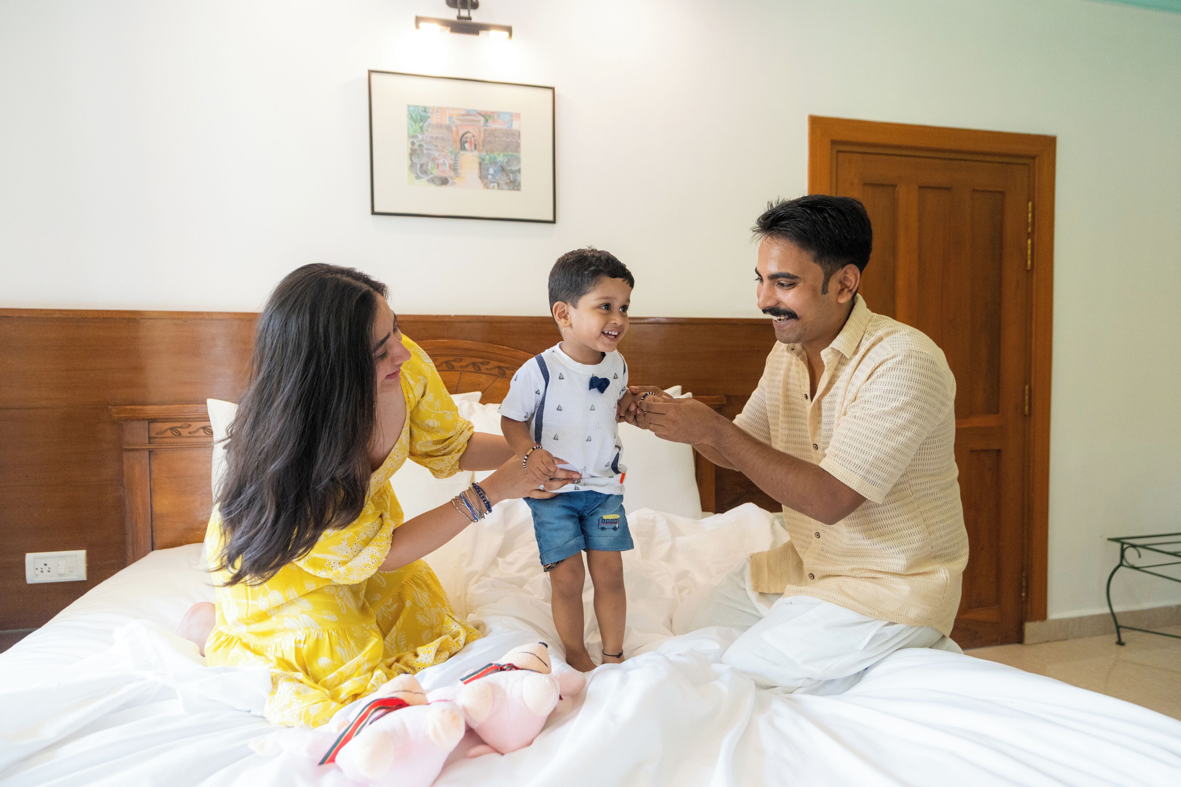 A family on a bed with a wooden headboard, a framed picture on the wall, and a door in the background.