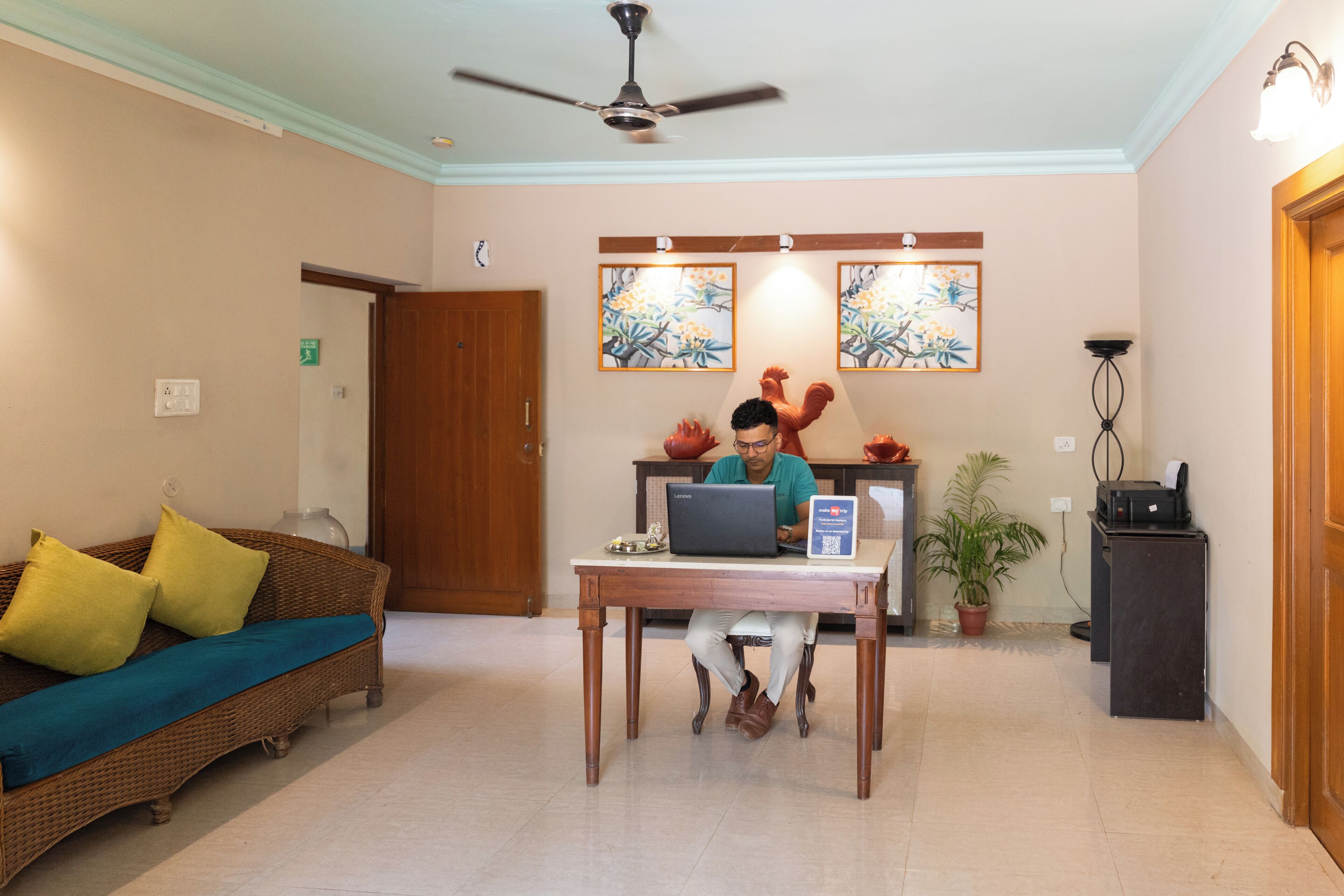 A reception area with a sofa, a table with a laptop, a ceiling fan, and two framed artworks on the wall.
