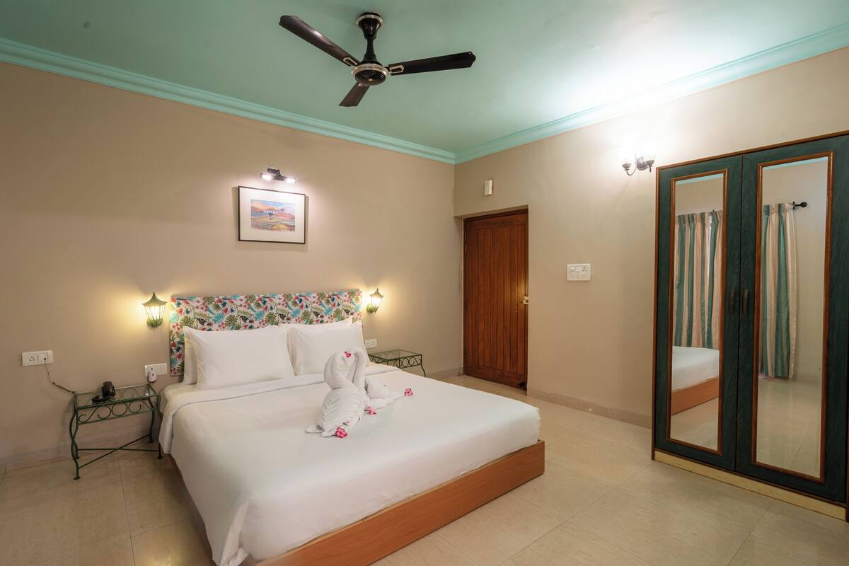 A hotel room with a bed, a ceiling fan, a nightstand, a wall-mounted light, a door, and a mirror.