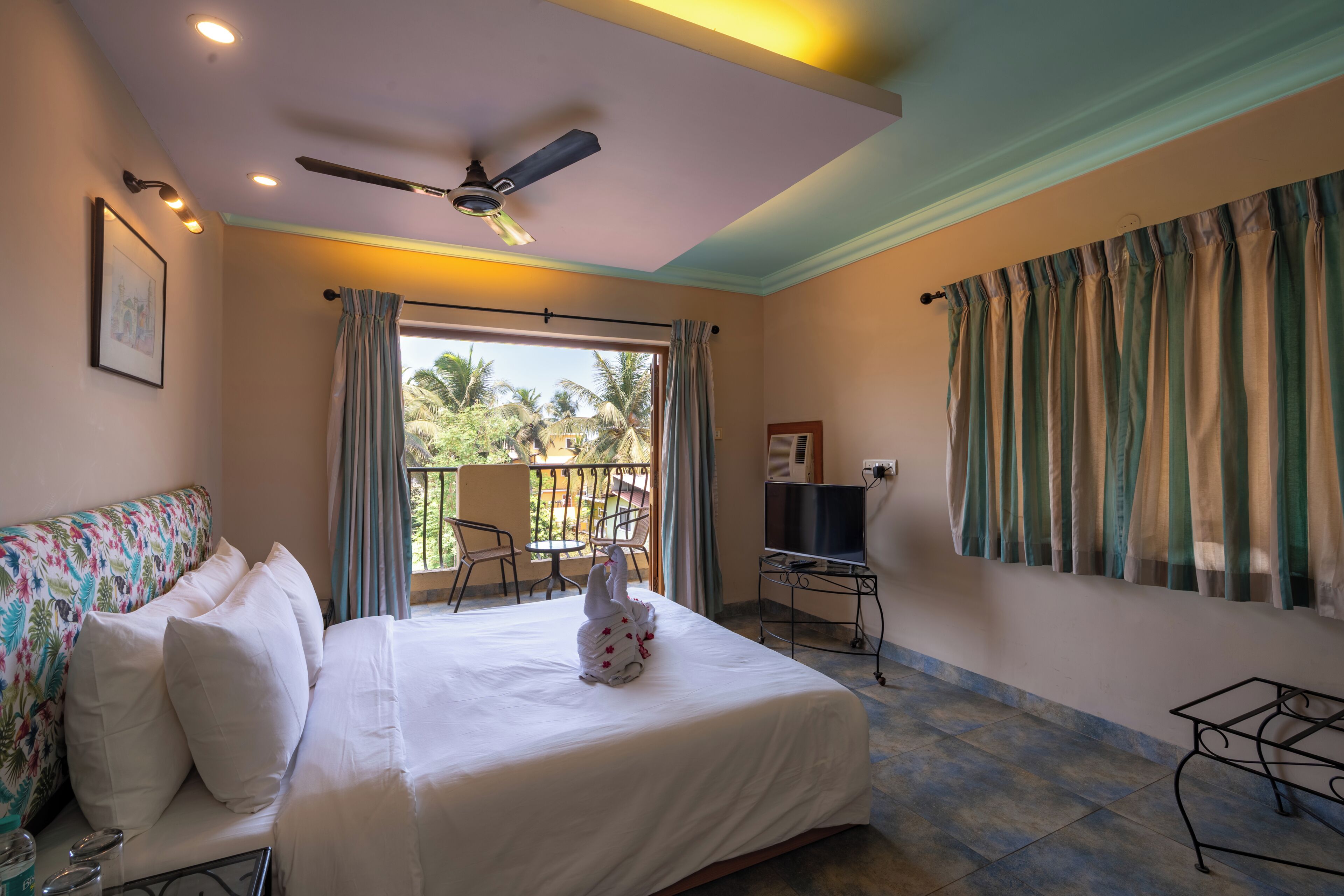 A hotel room with a bed, a ceiling fan, a TV, a balcony with a chair, and a view of palm trees.