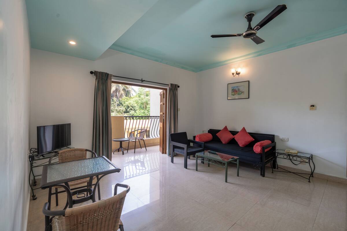 A living area with a sofa, a television, a dining table with chairs, a ceiling fan, and a balcony with a view of greenery.