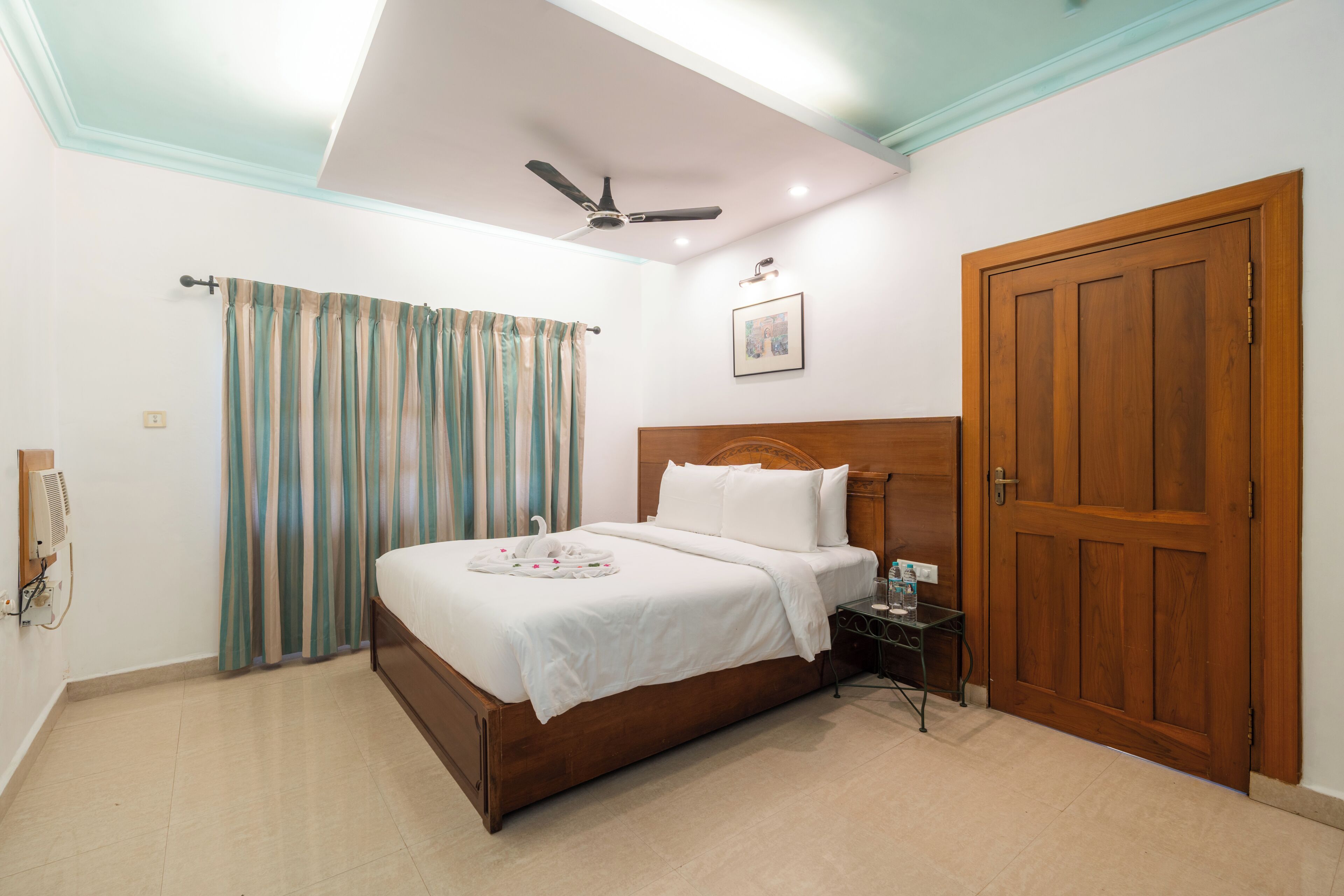 Um quarto com cama, ventilador de teto, porta de madeira e mesa de cabeceira.
