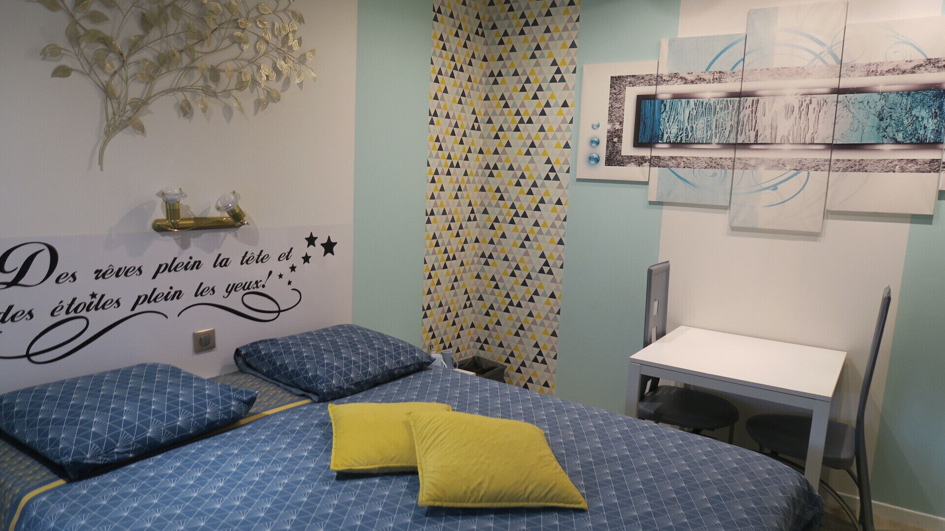 Un dormitorio con cama, escritorio y silla. Hay una pared decorativa con patrón geométrico y un adhesivo mural con texto.