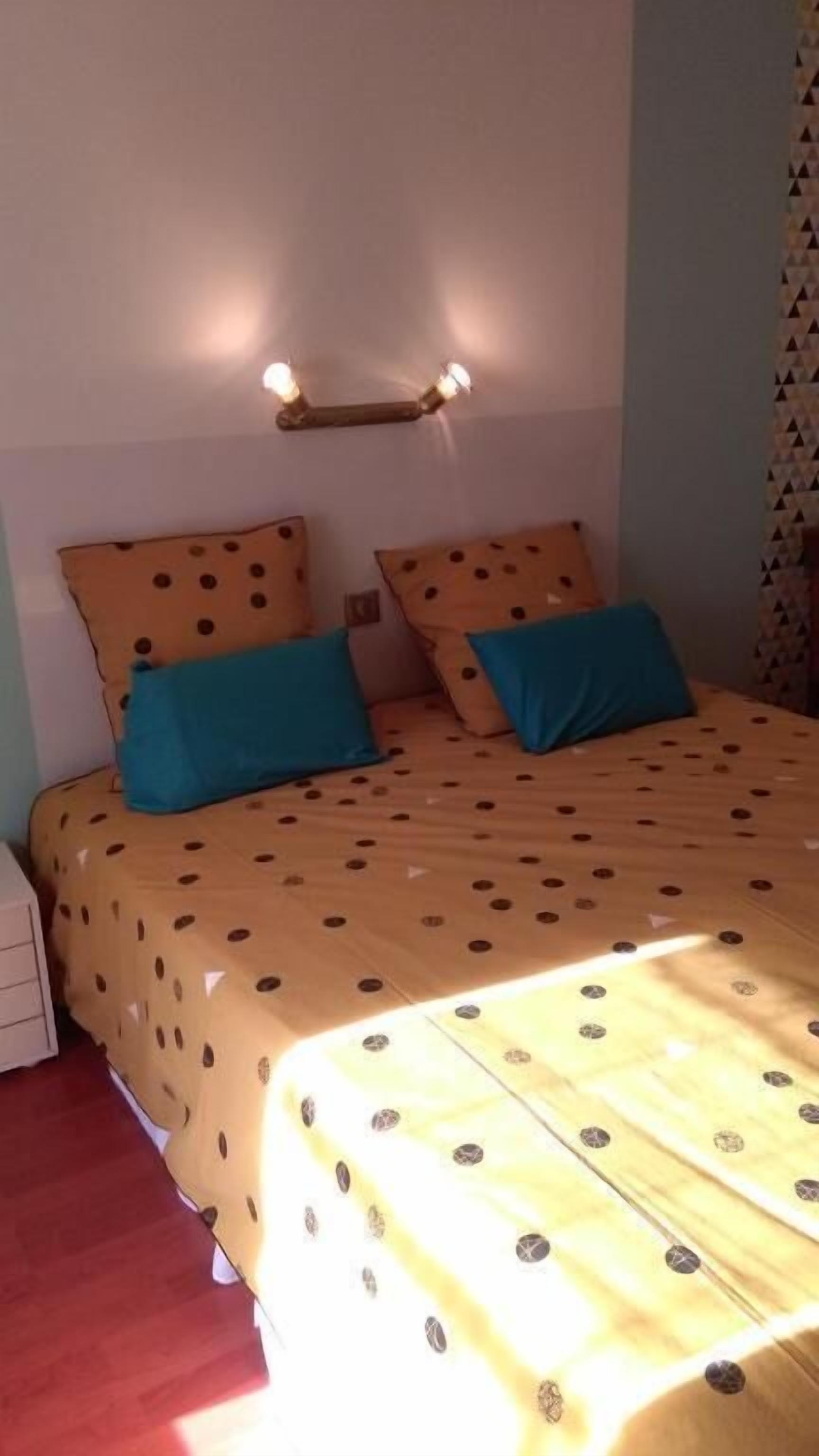 Cama com colcha de bolinhas, duas almofadas e luminária fixada na parede.