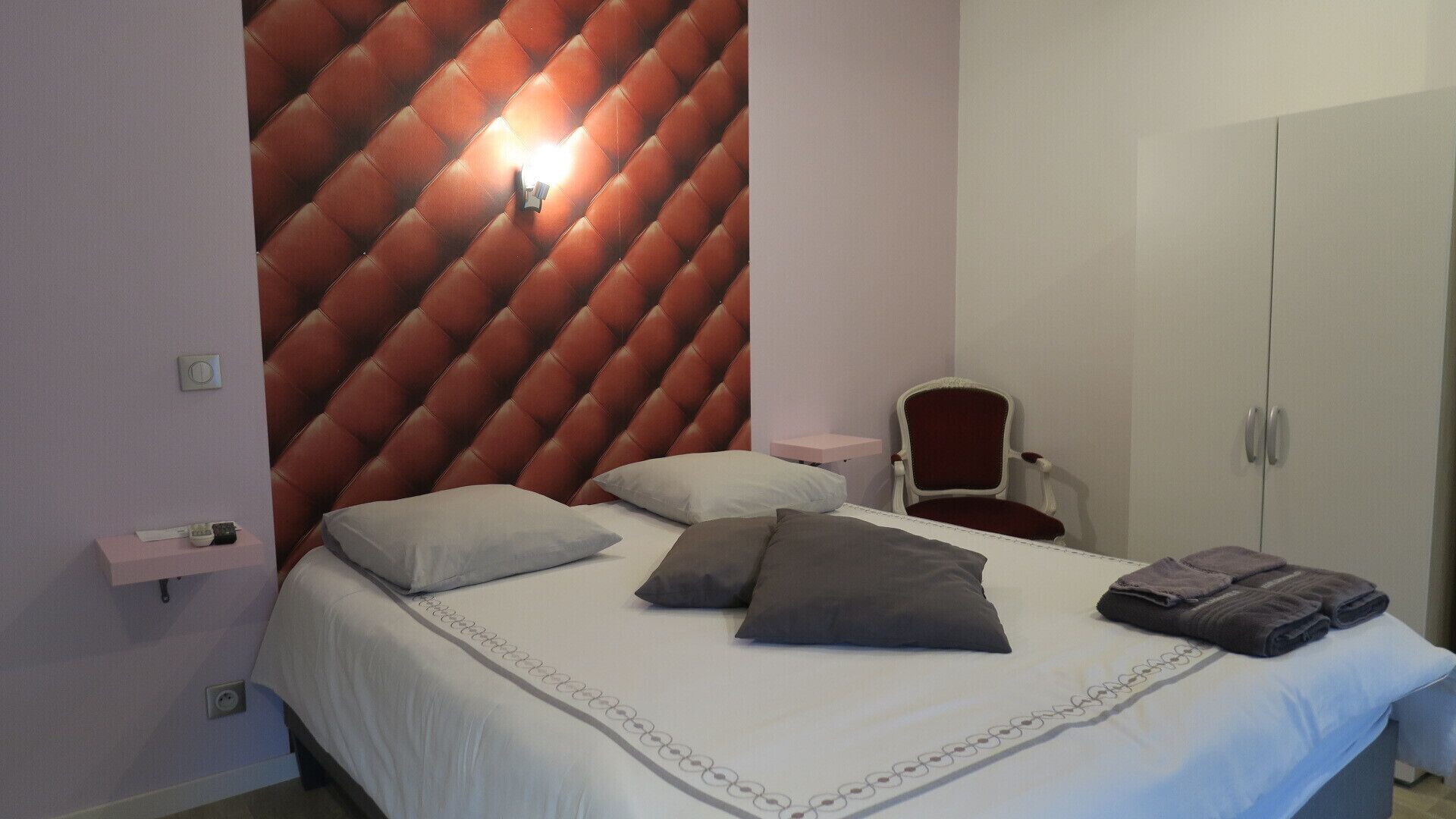 Une chambre à coucher avec une tête de lit capitonnée, un lit avec des oreillers, une chaise, une armoire et une table de chevet.