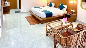 A hotel room with a bed, a ceiling fan, a nightstand, a bedside table, a chair, and wall-mounted artwork. - Devora Resort (Rishikesh)