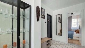 A modern room with a glass cabinet displaying glasses and candles, a wooden bench, a mirror, and a bed with a blue blanket. - Comfortable Apartment in Gdansk | Balcony | Workspace (Gdańsk)