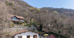 Ein zweistöckiges Haus mit einer Steinschmalseite, einem Balkon und Blick auf einen bewaldeten Hügel.