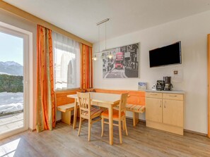Une pièce chaleureuse avec une table à manger en bois, deux chaises, un banc, un coin cuisine équipé d’une machine à café et un téléviseur à écran plat fixé au mur.
