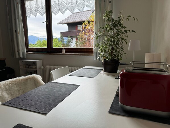 Un coin repas avec une table, des chaises, une plante en pot, un grille-pain rouge et une vue sur un immeuble par la fenêtre.