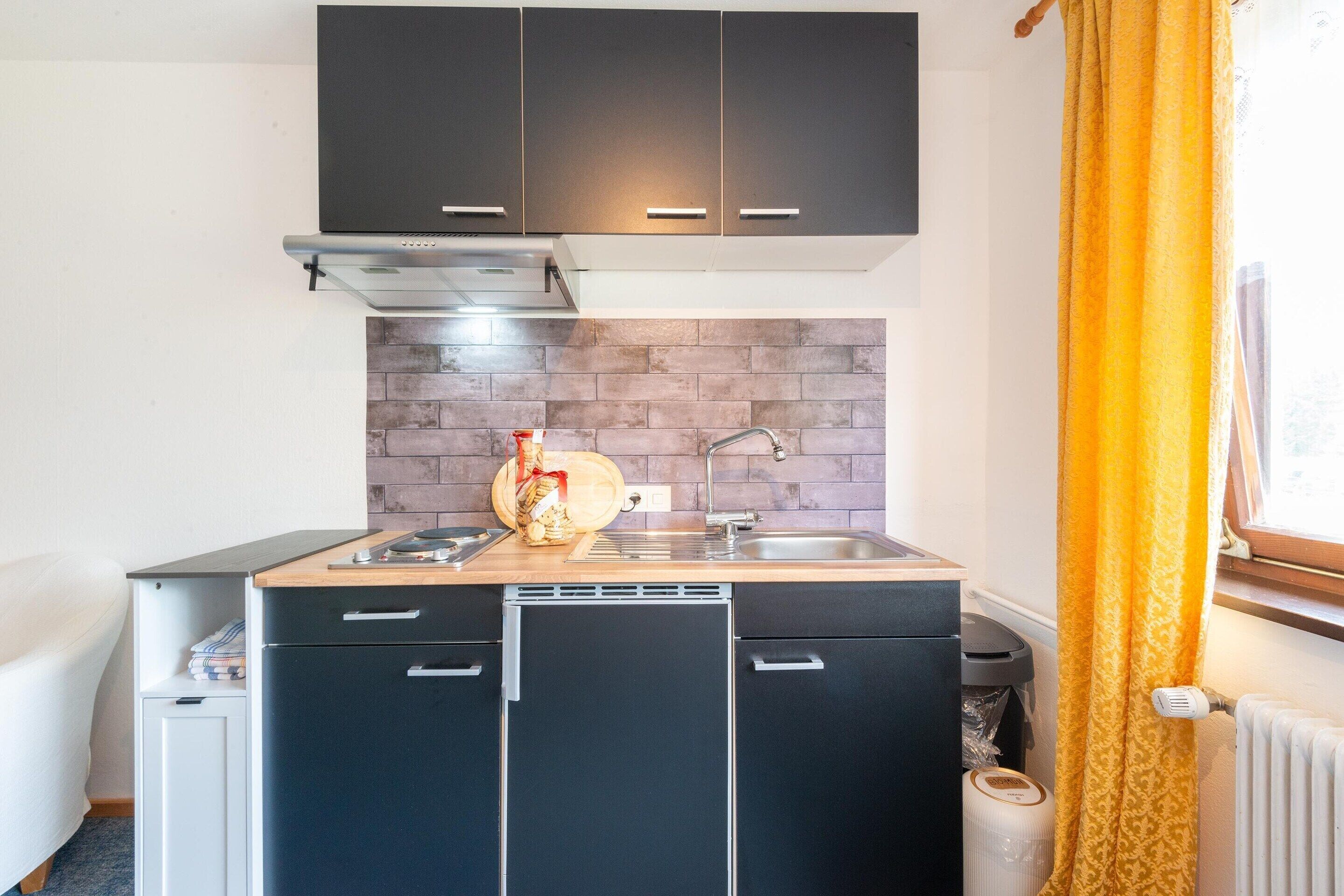 Une cuisine de taille réduite avec des meubles de couleur sombre, un plan de travail en bois, une cuisinière encastrée et une fenêtre ornée de rideaux jaunes.