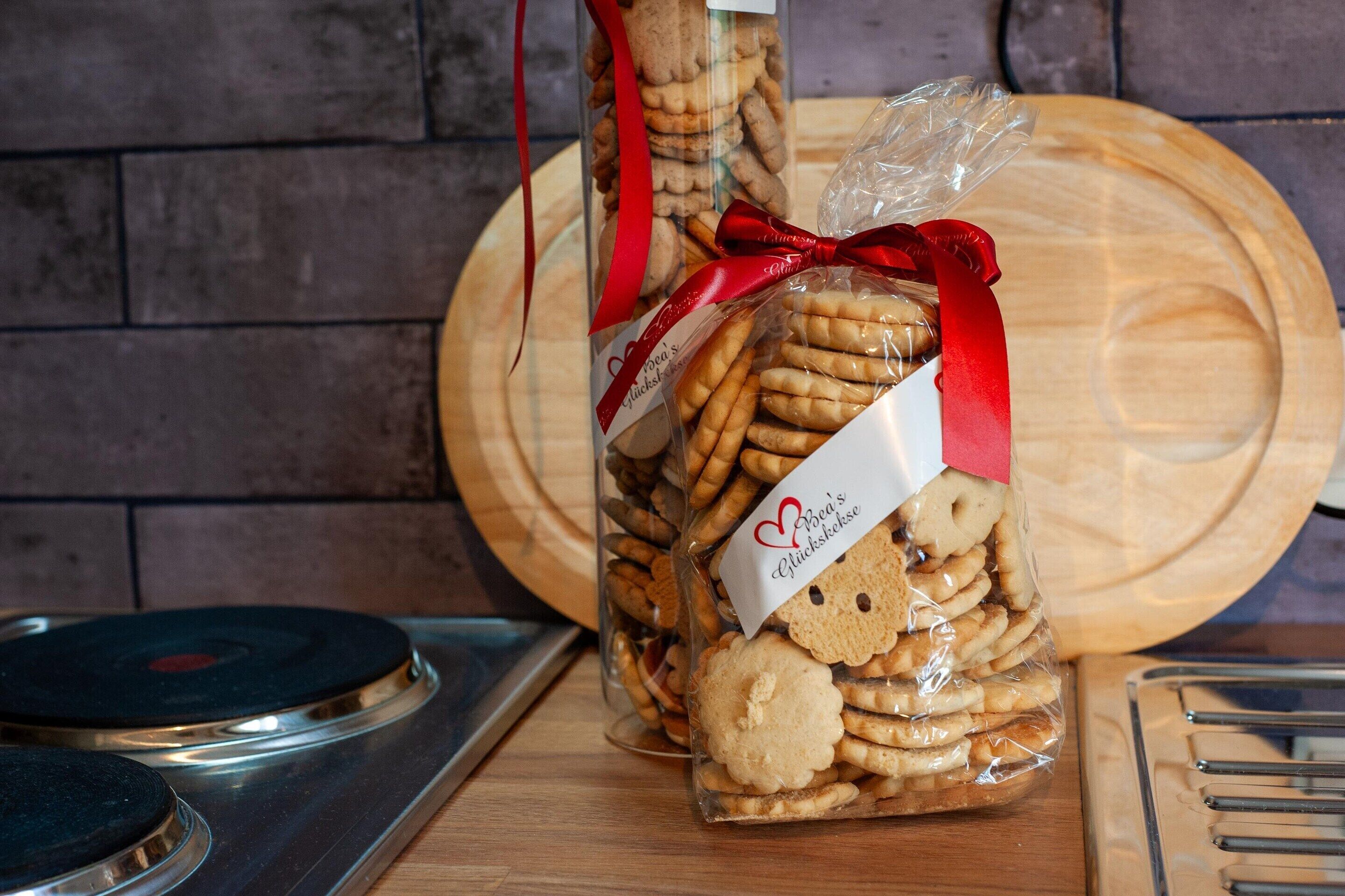 Un barattolo di vetro pieno di biscotti, decorato con un nastro rosso e un'etichetta, su una superficie di legno con un vassoio di metallo e un tagliere in secondo piano.