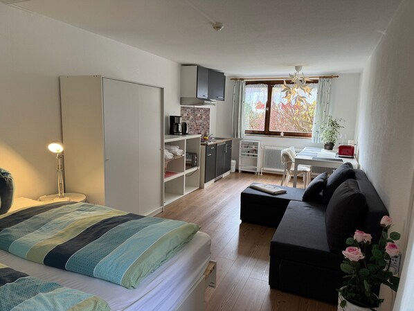 Une chambre comprenant un lit, une lampe de chevet, une armoire, un coin cuisine et un espace détente avec un canapé et une table.