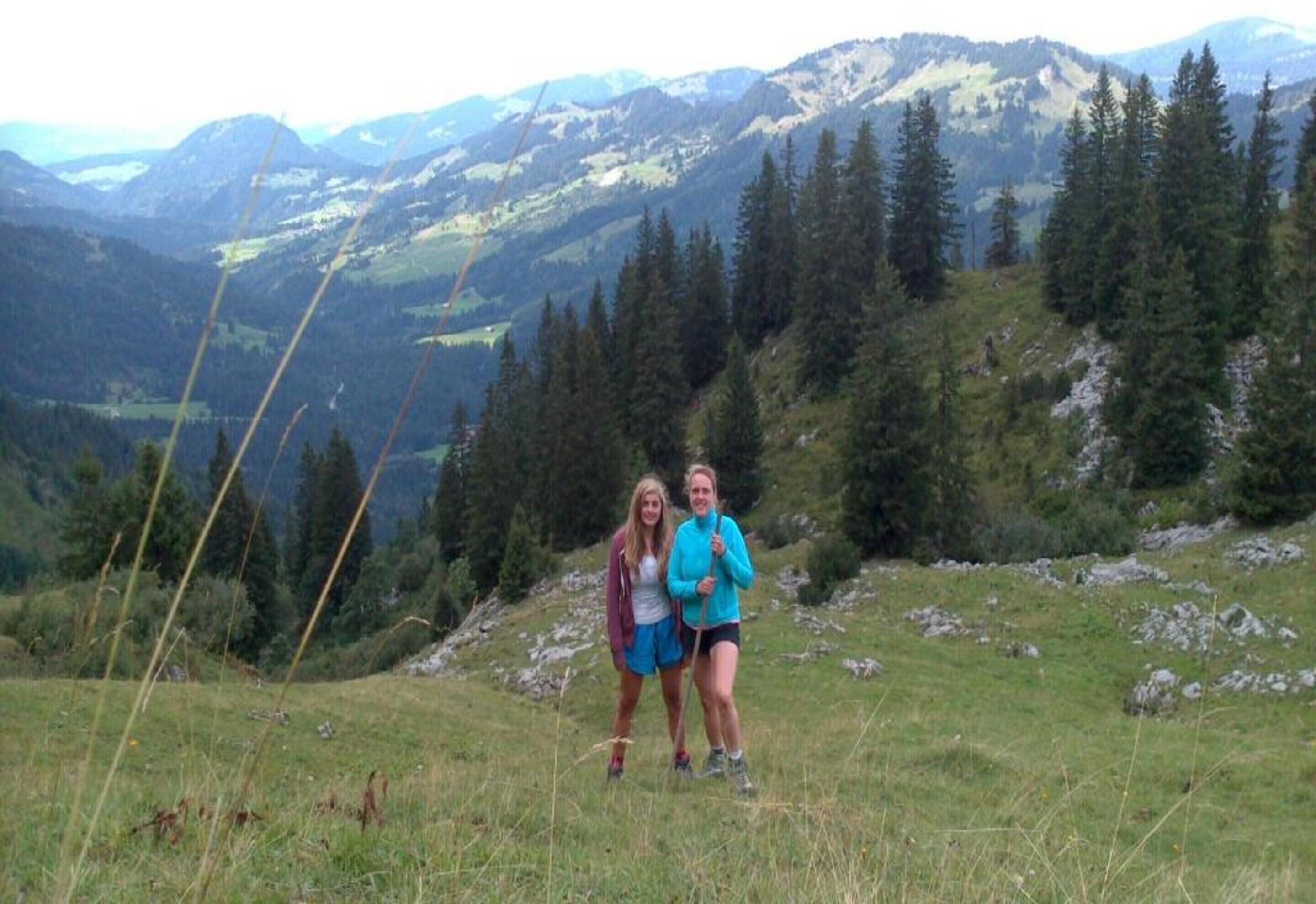 Dos personas en una colina cubierta de hierba, con un paisaje montañoso al fondo.