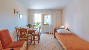 A small, well-lit room with a dining table, two wooden chairs, a single bed with a pillow, and a sofa.