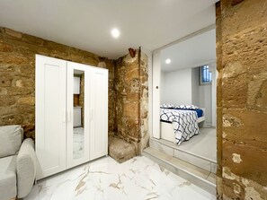 Een slaapkamer met stenen muren, een bed met beddengoed met een patroon, een witte garderobe en een badkamer zichtbaar door een geopende deur.
