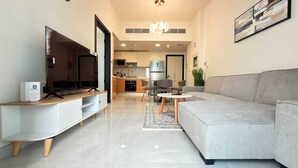 A modern living room with a flat-screen TV, a sofa, a coffee table, and a kitchen in the background. - Skyz Lifestyle 1BR Residences at Arjan (Dubai)